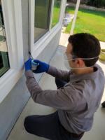 Man in a dust mask crouching next to exterior of a house while applying pesticide dust to a window frame.